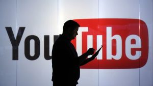 A man is seen as a silhouette as he checks a mobile device whilst standing against an illuminated wall bearing YouTube logo
